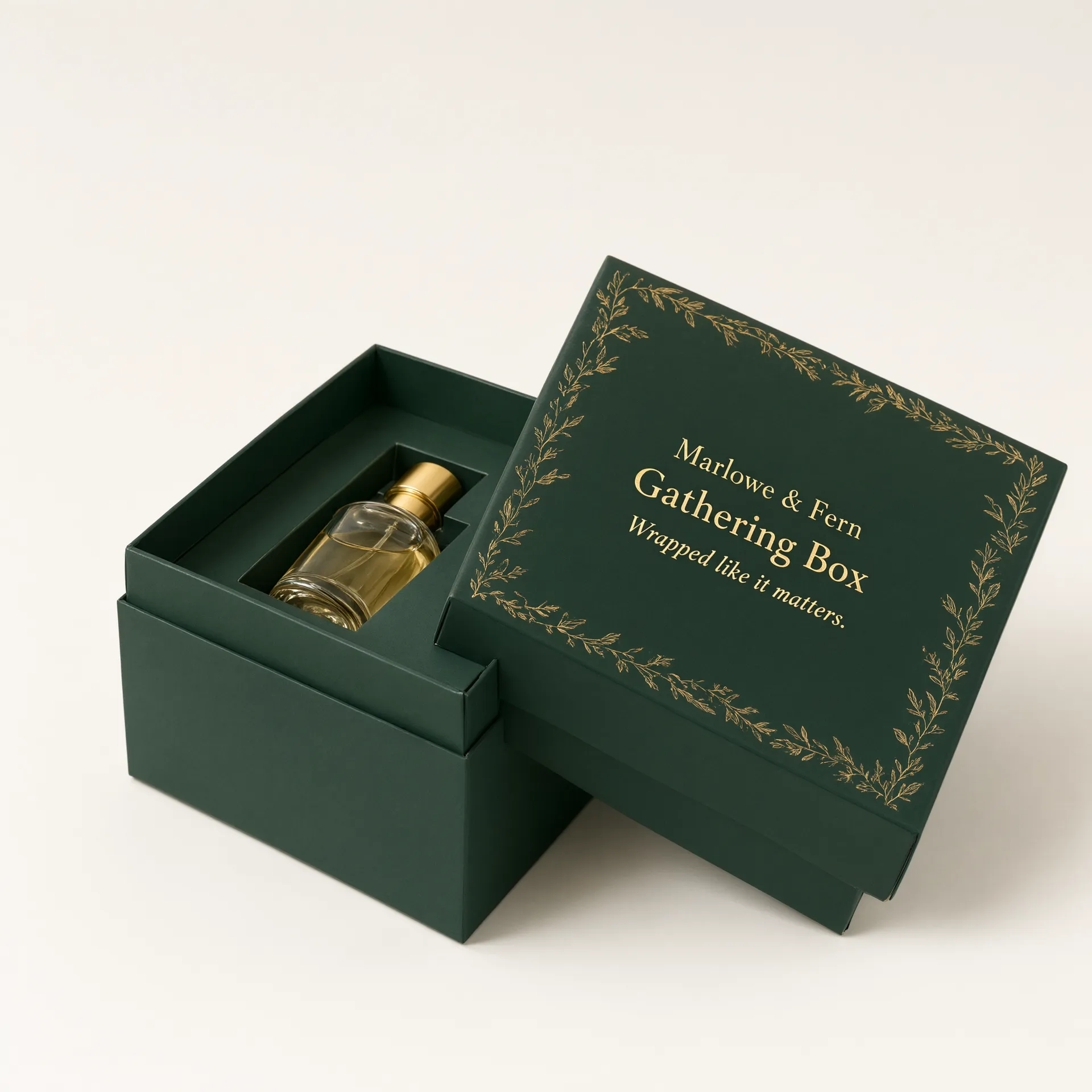 Open forest-green rigid gift box with gold foil botanical motif beside a fragrance bottle with a gold cap.